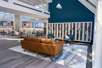 Photo of renovated Birkelund Lounge at the NHC. Includes new, white bookshelves, a brown leather couch,, brown coffee table, and blue fabric chairs on a color coordinating rug. Features a dark blue accent wall